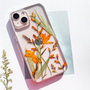 May include: A clear phone case with pressed orange and green flowers. The case is designed for an iPhone with a camera lens visible.