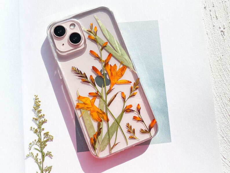 May include: A clear phone case with pressed orange and green flowers. The case is designed for an iPhone with a camera lens visible.
