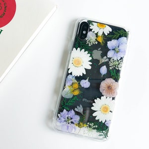 May include: A clear phone case with a floral design. The case features pressed flowers in shades of white, purple, and green. The text "iPhone" is visible on the back of the phone.
