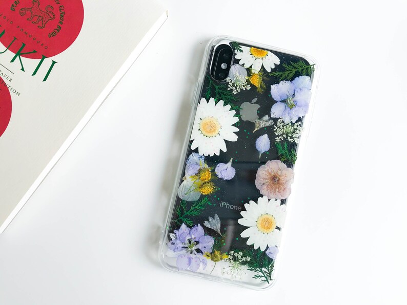 May include: A clear phone case with a floral design. The case features pressed flowers in shades of white, purple, and green. The text "iPhone" is visible on the back of the phone.