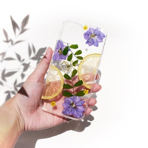 Puede incluir: Una funda transparente para tel&eacute;fono con flores prensadas y rodajas de lim&oacute;n. Las flores son moradas y amarillas, y las rodajas de lim&oacute;n son de color amarillo claro. La funda se sostiene en una mano.