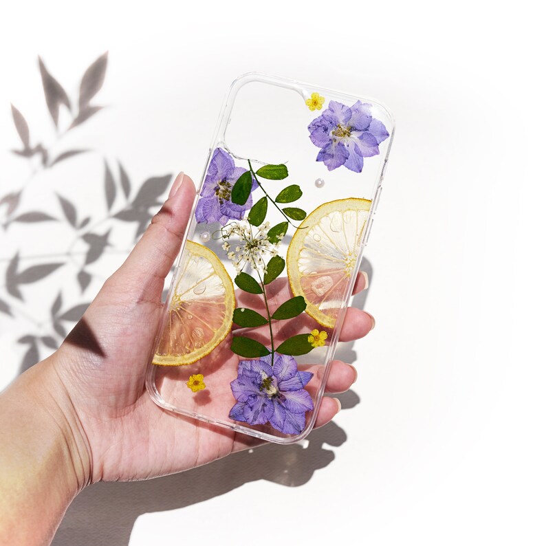 Puede incluir: Una funda transparente para tel&eacute;fono con flores prensadas y rodajas de lim&oacute;n. Las flores son moradas y amarillas, y las rodajas de lim&oacute;n son de color amarillo claro. La funda se sostiene en una mano.