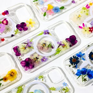 Peut inclure: Coques de téléphone transparentes avec des fleurs séchées de différentes couleurs, notamment violet, jaune et bleu. Chaque coque présente un motif circulaire blanc. Les coques sont disposées sur une surface blanche. Le texte "Kristin ABBLOOM Floral Design" est visible.