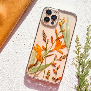 May include: A clear phone case with a pressed flower design. The case features orange and green flowers with a light pink background. The case is designed for an iPhone with a camera bump.