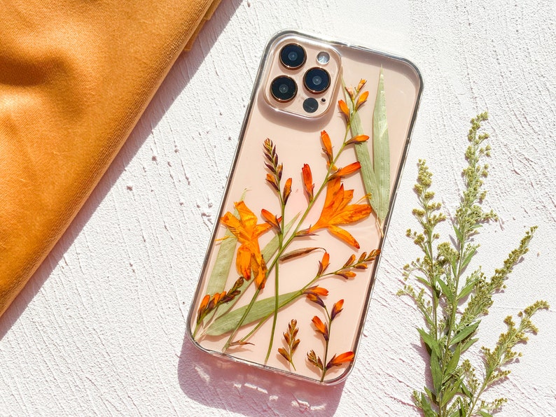 May include: A clear phone case with a pressed flower design. The case features orange and green flowers with a light pink background. The case is designed for an iPhone with a camera bump.