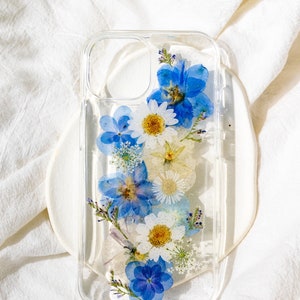 May include: A clear phone case with a design of pressed blue and white flowers. The case includes glitter. The text "STYLE F" is visible at the top of the image. The text "This case includes glitter. Note 'no glitter' at checkout if preferred." is visible at the bottom of the image.