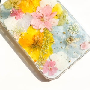 May include: A clear phone case with pressed flowers and gold leaf accents. The flowers are yellow, pink, blue, and white.