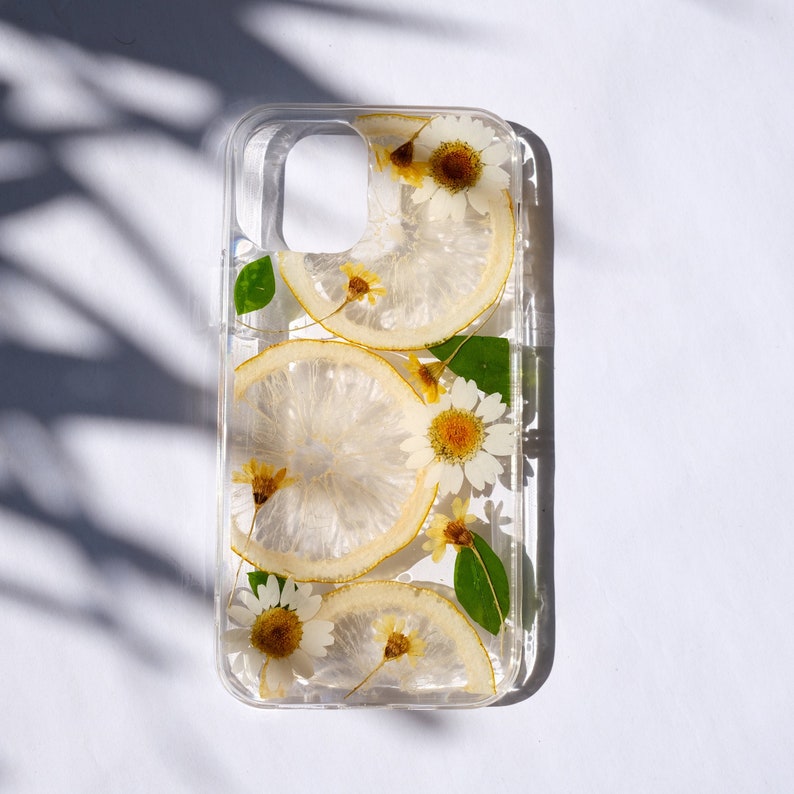 Puede incluir: Una funda transparente para tel&eacute;fono con un dise&ntilde;o floral. La funda presenta flores prensadas y rodajas de lim&oacute;n en una resina transparente. Las flores son blancas y amarillas, y las rodajas de lim&oacute;n son de un amarillo p&aacute;lido.