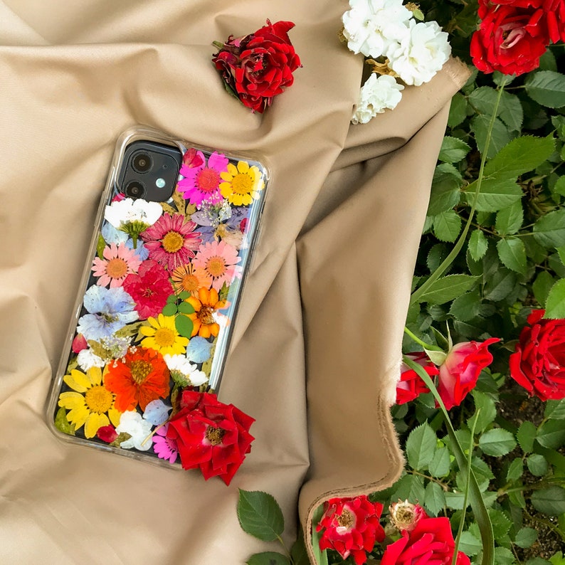 May include: A clear phone case with a pressed flower design. The case features a variety of colorful flowers, including pink, yellow, red, and orange. The case is lying on a brown fabric surface.