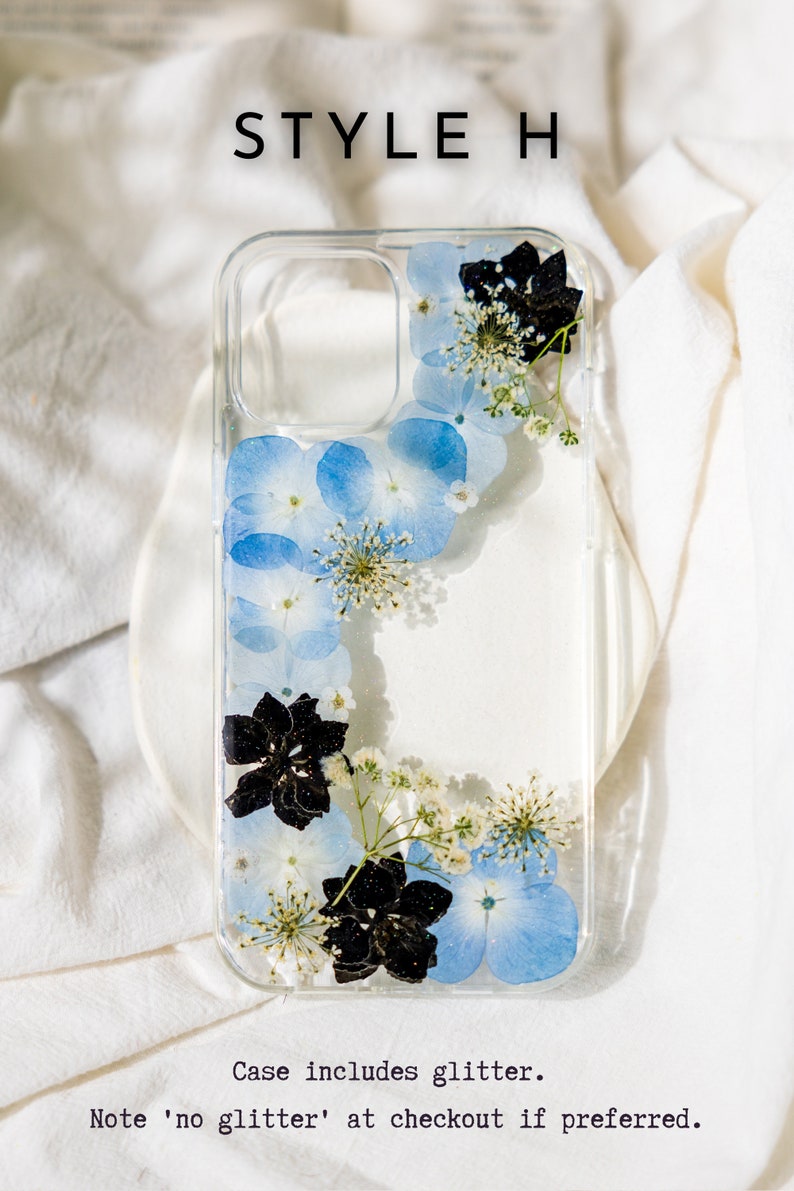 May include: A clear phone case with a design of pressed blue and white flowers and black flowers. The case includes glitter.
