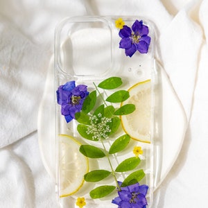 May include: A clear phone case with a pressed flower design. The case features purple flowers, green leaves, and a lemon slice. The text "STYLE A" is at the top of the image. The text "This case includes pearls. Note 'no pearls' at checkout if preferred." is at the bottom of the image.
