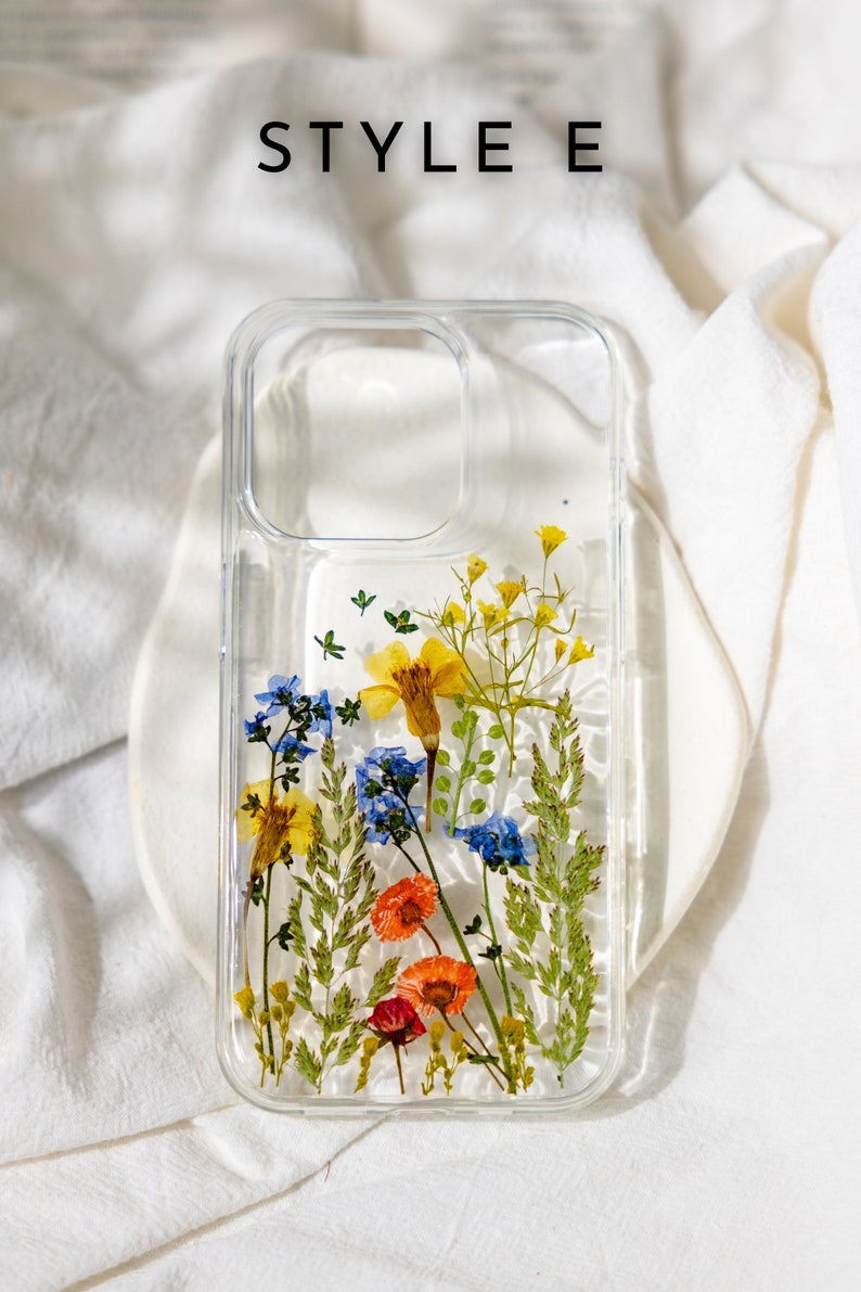 May include: A clear phone case with pressed flowers inside. The flowers are yellow, blue, orange, and red. The case has a raised lip around the camera lens.
