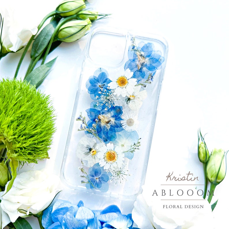 May include: A clear phone case with pressed blue and white flowers. The case has a white background and the flowers are arranged vertically. The text "Kristin ABLOOM FLORAL DESIGN" is printed on the bottom of the case.