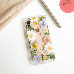 May include: A clear phone case with a floral design. The case features pressed flowers in shades of white, yellow, pink, and purple. The flowers are arranged in a random pattern and are visible through the clear case. The case is designed for an iPhone.