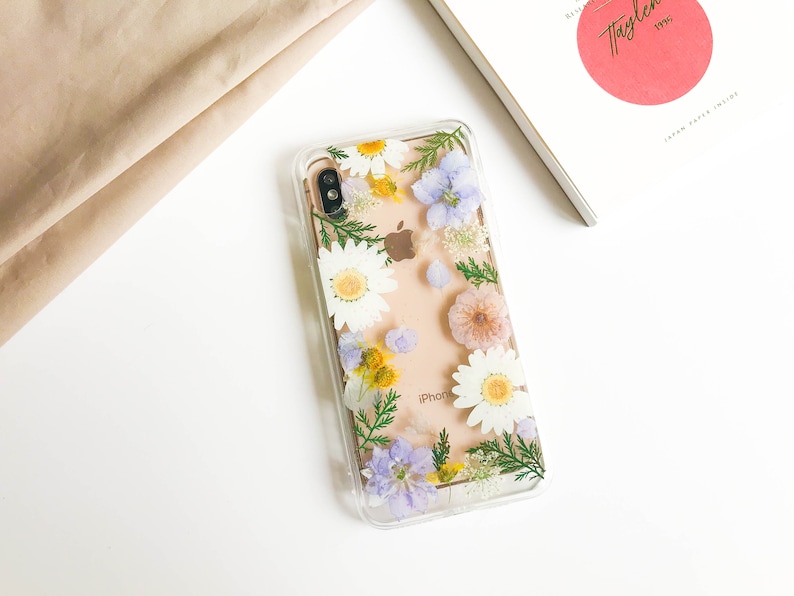 May include: A clear phone case with a floral design. The case features pressed flowers in shades of white, yellow, pink, and purple. The flowers are arranged in a random pattern and are visible through the clear case. The case is designed for an iPhone.