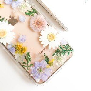 May include: A clear phone case with pressed flowers and greenery. The flowers are white, purple, and yellow. The case has the word "phone" printed on it.