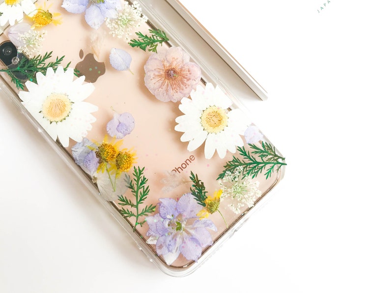 May include: A clear phone case with pressed flowers and greenery. The flowers are white, purple, and yellow. The case has the word "phone" printed on it.