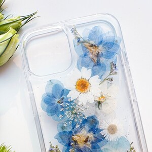 May include: A clear phone case with pressed blue and white flowers. The flowers are arranged in a cluster and appear to be pressed between two layers of clear plastic.