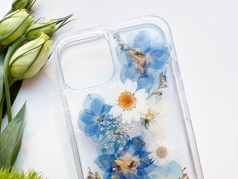 May include: A clear phone case with pressed blue and white flowers. The flowers are arranged in a cluster and appear to be pressed between two layers of clear plastic.