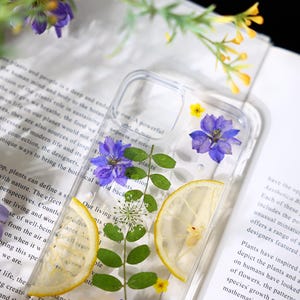 Puede incluir: Una funda transparente para tel&eacute;fono con flores prensadas y una rodaja de lim&oacute;n en su interior. Las flores son moradas y azules, y la rodaja de lim&oacute;n es amarilla. La funda est&aacute; sobre un libro blanco con texto.