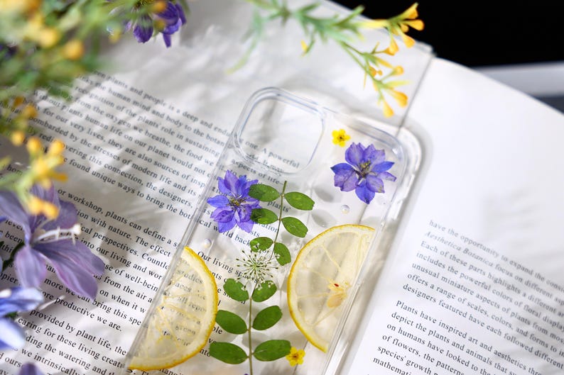 Puede incluir: Una funda transparente para tel&eacute;fono con flores prensadas y una rodaja de lim&oacute;n en su interior. Las flores son moradas y azules, y la rodaja de lim&oacute;n es amarilla. La funda est&aacute; sobre un libro blanco con texto.