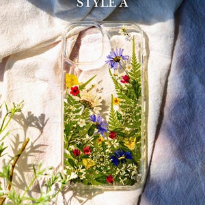 May include: A clear phone case with pressed flowers and greenery inside. The flowers are in shades of blue, yellow, red, and white. The case has a clear back and sides, and the flowers are visible through the case. The case is labeled "STYLE A".