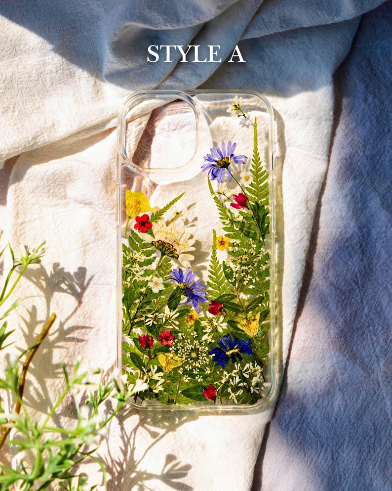 May include: A clear phone case with pressed flowers and greenery inside. The flowers are in shades of blue, yellow, red, and white. The case has a clear back and sides, and the flowers are visible through the case. The case is labeled "STYLE A".