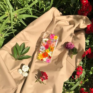May include: A clear phone case with a pressed flower design. The case features a variety of colorful flowers, including pink, yellow, orange, and white.