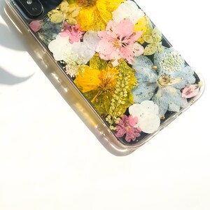 May include: A clear phone case with a pressed flower design. The flowers are yellow, pink, blue, and white. The case has a black background.