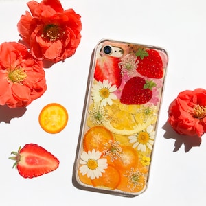 May include: A clear phone case with a pressed flower and fruit design. The case features dried strawberries, lemons, and orange slices, as well as white flowers. The case is surrounded by red flowers.