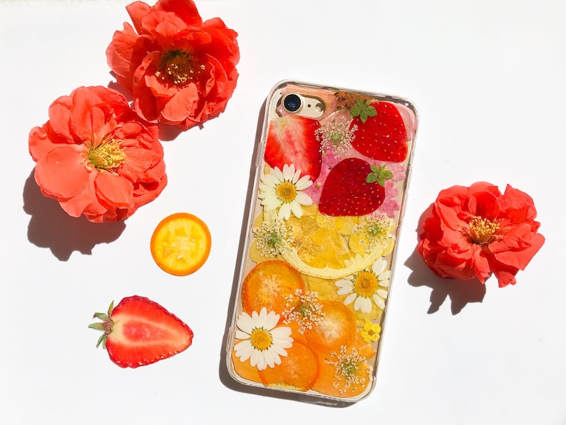May include: A clear phone case with a pressed flower and fruit design. The case features dried strawberries, lemons, and orange slices, as well as white flowers. The case is surrounded by red flowers.