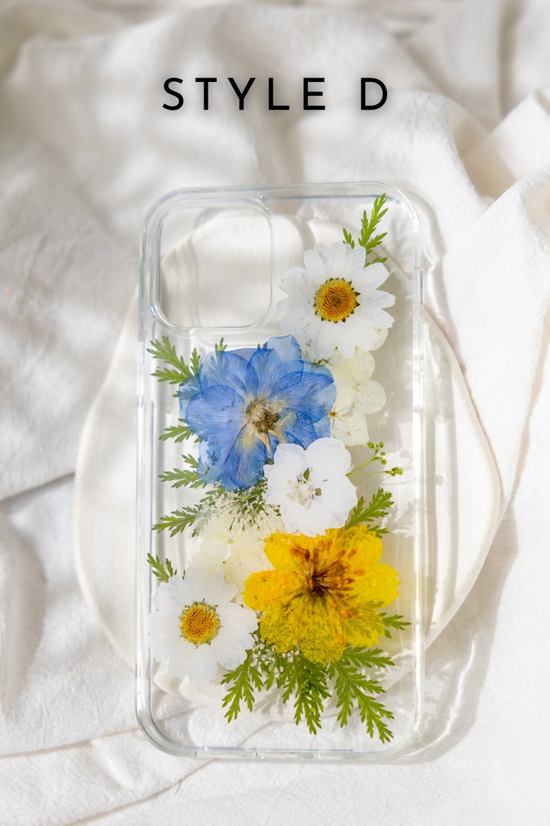May include: A clear phone case with pressed flowers. The flowers are white, blue, and yellow. The case has a clear back and sides, and the flowers are visible through the case. The case is designed to fit a smartphone.
