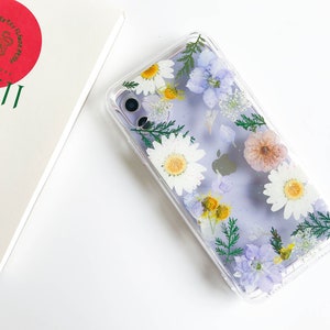 May include: A clear phone case with a pressed flower design. The case features white daisies, purple flowers, and green leaves. The case is designed to fit a smartphone.
