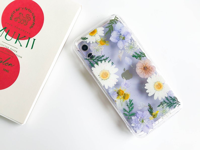May include: A clear phone case with a pressed flower design. The case features white daisies, purple flowers, and green leaves. The case is designed to fit a smartphone.