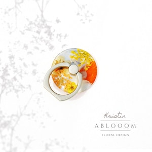 May include: A silver phone ring holder with a clear resin base filled with pressed flowers in shades of yellow, orange, and white. The ring holder is designed to be attached to the back of a phone.
