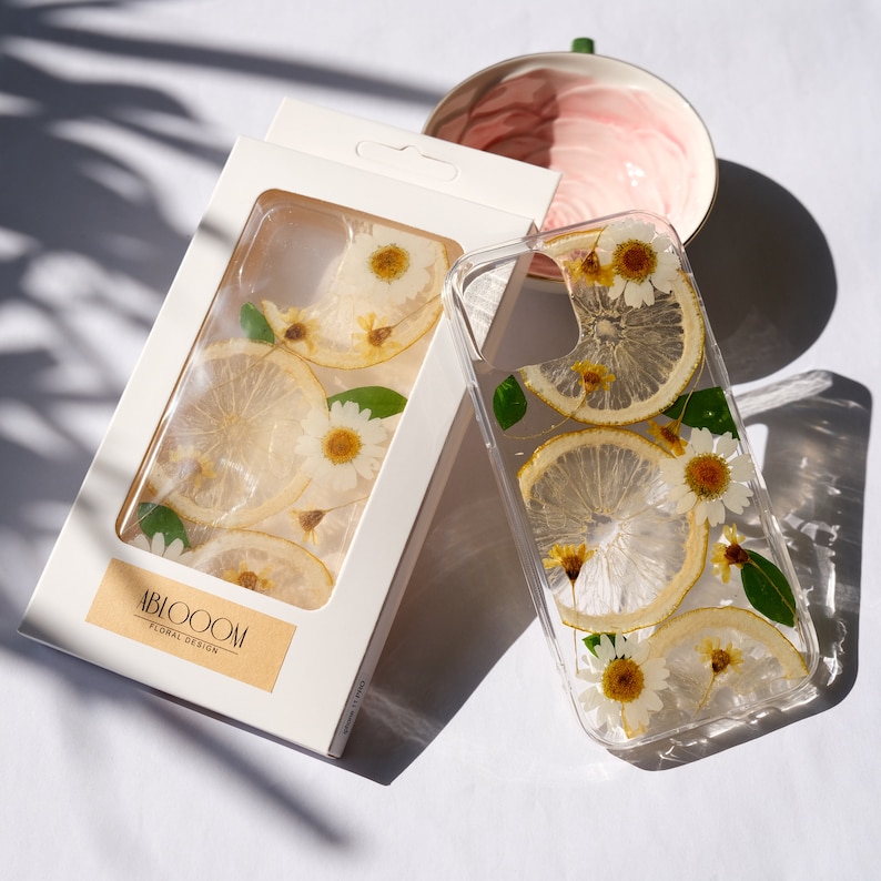 Puede incluir: Una funda transparente para tel&eacute;fono con un dise&ntilde;o floral que incluye rodajas de lim&oacute;n secas y margaritas blancas. La funda est&aacute; en una caja blanca con el texto "ABLOOM FLORAL DESIGN".