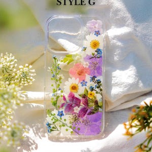 May include: A clear phone case with a floral design. The case is filled with pressed flowers in shades of pink, purple, blue, and yellow. The text "STYLE G" is printed on the top of the case.