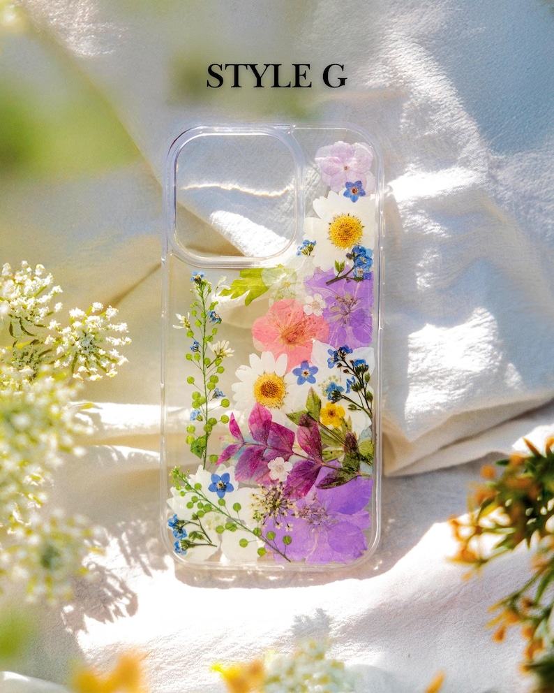 May include: A clear phone case with a floral design. The case is filled with pressed flowers in shades of pink, purple, blue, and yellow. The text "STYLE G" is printed on the top of the case.