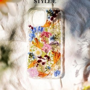May include: A clear phone case with a pressed flower design. The case features a variety of flowers in shades of blue, pink, orange, and white. The text "STYLE E" is printed on the top of the case.