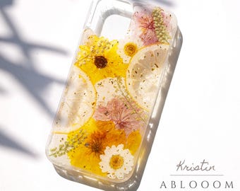 May include: A clear phone case with dried flowers and lemon slices embedded in resin. The flowers are yellow, pink, and white. The case has a gold glitter effect. The text "Kristin ABLOOOOM FLORAL DESIGN" is visible at the bottom of the image.