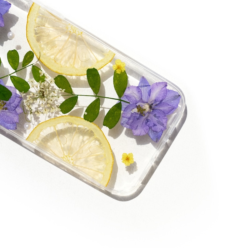 Puede incluir: Una funda transparente para tel&eacute;fono con flores prensadas y rodajas de lim&oacute;n. Las flores son moradas y amarillas, y las rodajas de lim&oacute;n son amarillas.