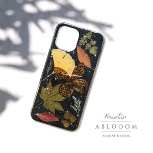 May include: Black phone case with a pressed flower design featuring a butterfly, leaves, and gold accents. The case has a clear background and the design is visible through the case. The text "Kristin ABLOOOOM FLORAL DESIGN" is visible in the bottom right corner of the image.