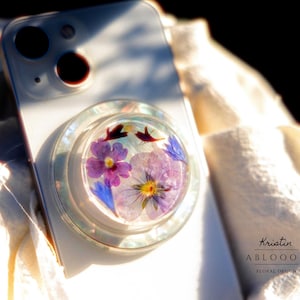 以下が含まれることがあります： 紫と黄色の花と青いアクセントが特徴のフローラルデザインの透明な電話グリップ。 電話グリップは白いスマートフォンに取り付けられています。 テキスト「Kristin ABLOOM Floral Design」が表示されます。