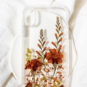 May include: A clear phone case with pressed orange and yellow flowers inside. The case is on a white surface. "STYLE G" is written above the case.