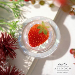 Puede incluir: Un tapón de botella redondo y transparente con una fresa roja vibrante incrustada. La fresa tiene hojas verdes y está colocada sobre un fondo iridiscente. El tapón forma parte de una colección de diseño floral.