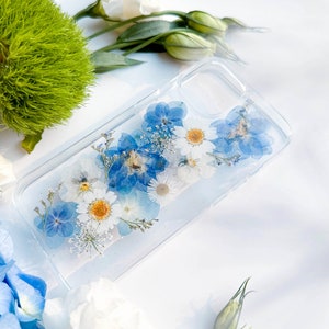 May include: A clear phone case with pressed blue and white flowers. The flowers are arranged in a random pattern and are visible through the clear case.