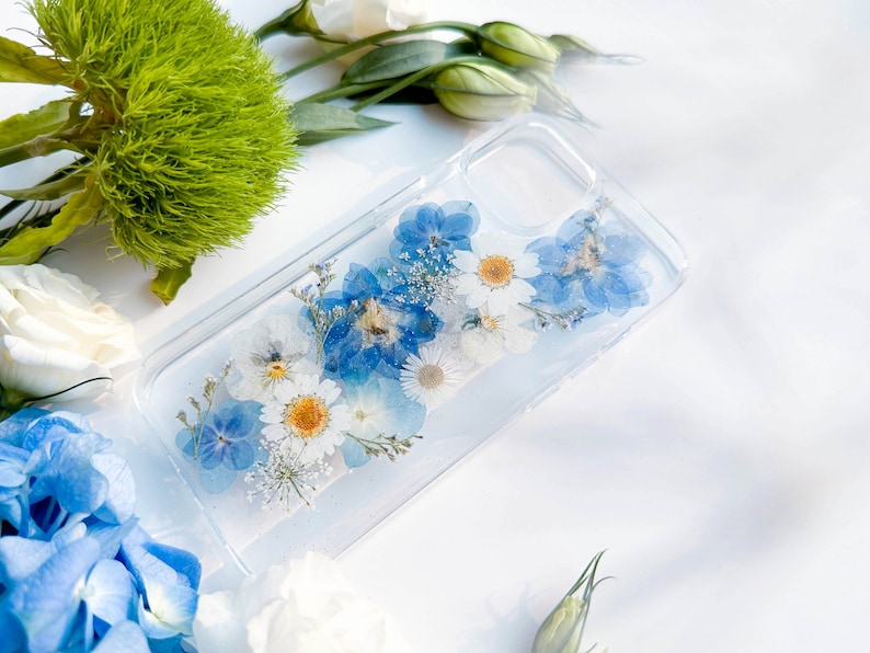 May include: A clear phone case with pressed blue and white flowers. The flowers are arranged in a random pattern and are visible through the clear case.