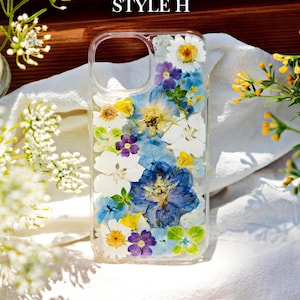 May include: A clear phone case with a pressed flower design. The case features a variety of blue, white, yellow, and purple flowers. The text "STYLE H" is visible at the top of the image.