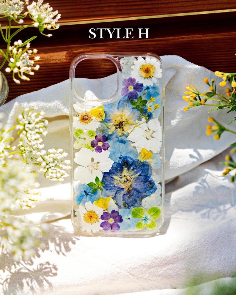 May include: A clear phone case with a pressed flower design. The case features a variety of blue, white, yellow, and purple flowers. The text "STYLE H" is visible at the top of the image.