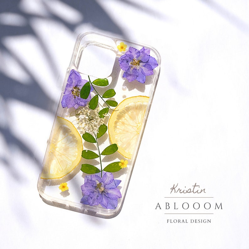 Puede incluir: Una funda transparente para tel&eacute;fono con flores y rodajas de lim&oacute;n prensadas. Las flores son moradas y amarillas, y los limones son amarillos. La funda tiene el texto "Kristin ABLOOOOM FLORAL DESIGN" en la parte posterior.